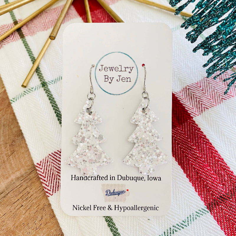 A pair of white glitter Christmas tree earrings displayed on a card with 'Jewelry by Jen' written on it, placed on a red and green placemat with gold decorations.