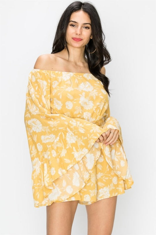 A mustard yellow bell sleeve romper with a floral print, featuring a belted elastic waistband and shorts lined within. The garment is draped over a mannequin, showcasing the fit and design.